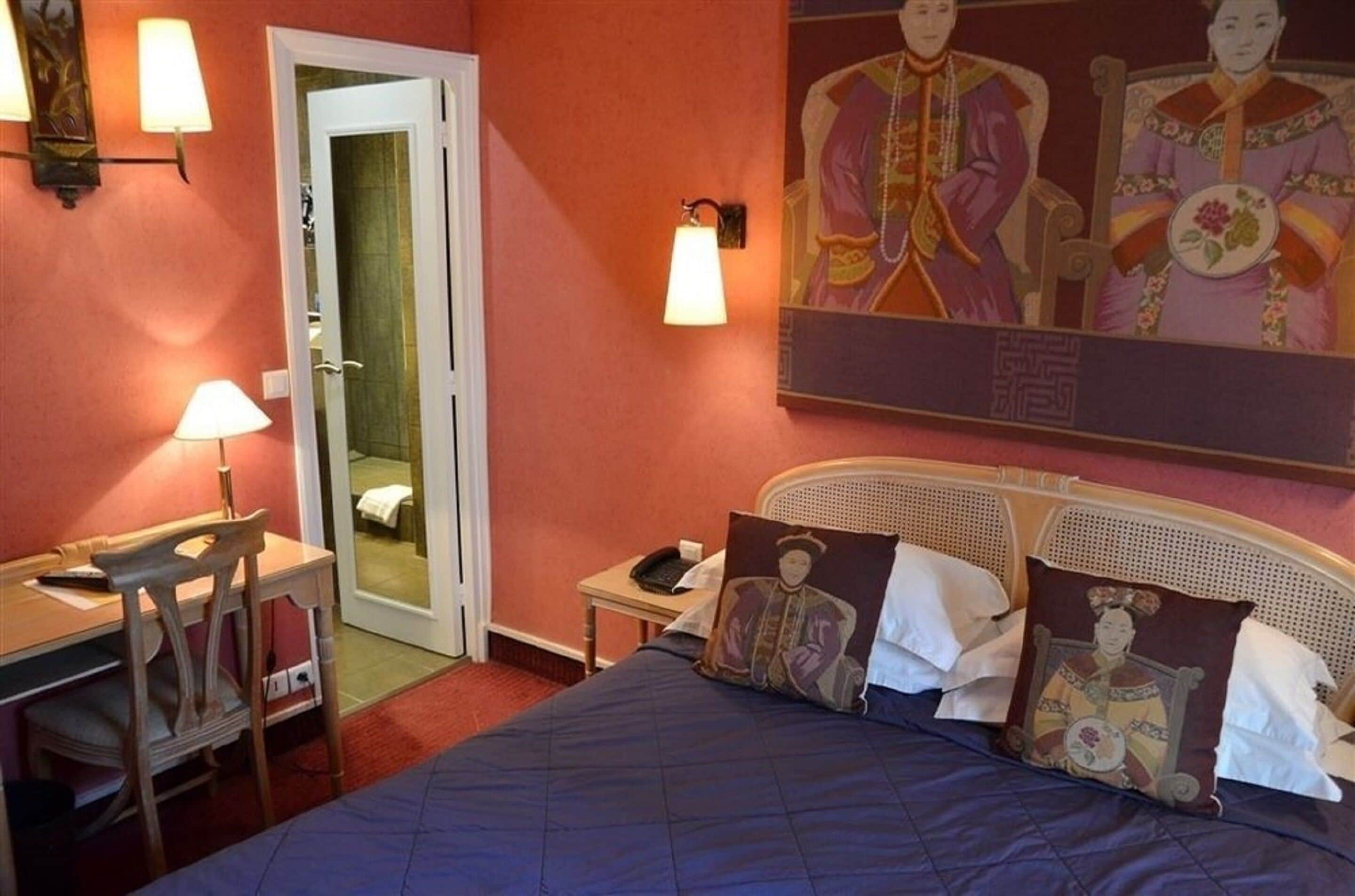 A hotel room with a bed, desk, chair, and a painting on the wall.