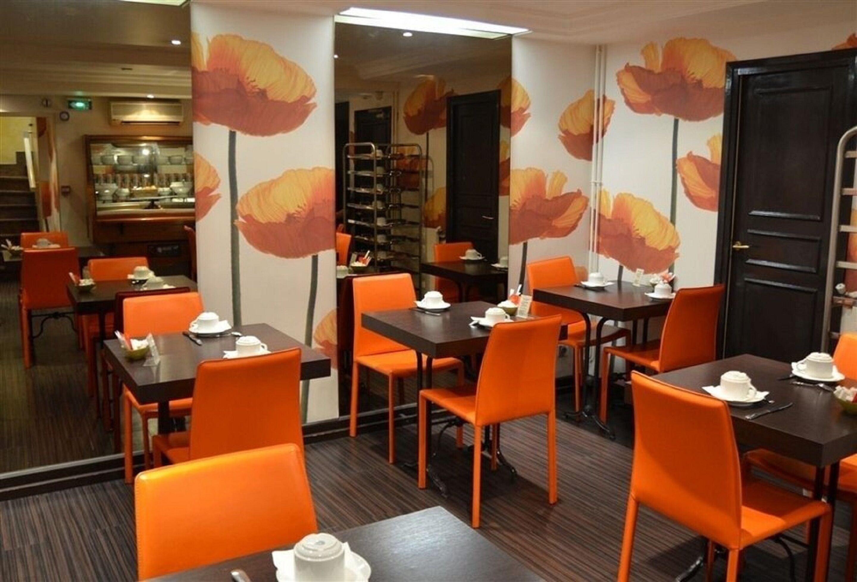 Un coin repas avec des chaises et des tables orange, une cuisine en arrière-plan et un mur orné d’un motif floral.