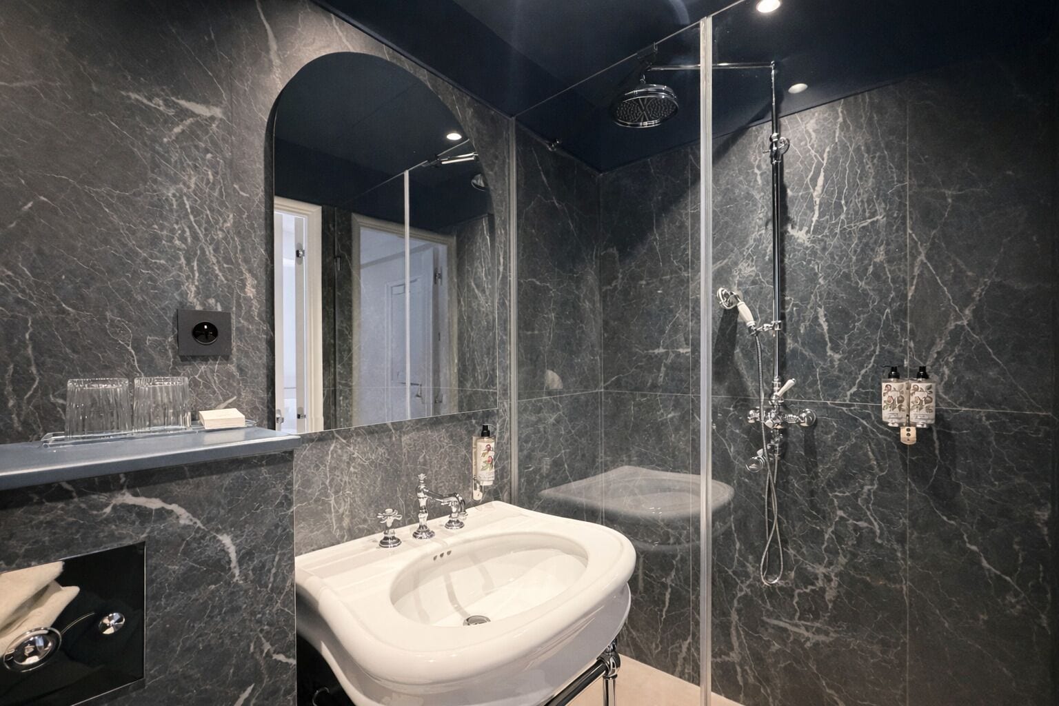 Ein modernes Badezimmer mit Marmorwand, einer in Glas eingefassten Dusche, einem weißen Waschbecken und einem Spiegel.