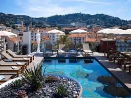 A rooftop pool area with lounge chairs and umbrellas, surrounded by buildings and a view of a hill.