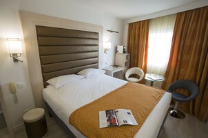 Hotellihuone, jossa on suuri sänky, yöpöytä, tuoli, pieni pöytä, lamppu ja roskakori.