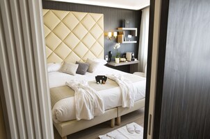 A modern hotel room with a large bed, a bedside table with a lamp, and a small shelf with a book and a phone.