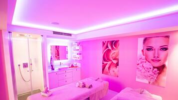 Une salle de spa aux couleurs rose, comprenant deux tables de massage, une coiffeuse avec un miroir et un espace douche.