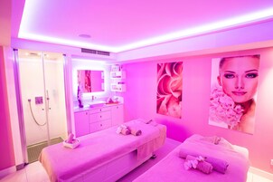 Une salle de spa aux couleurs rose, comprenant deux tables de massage, une coiffeuse avec un miroir et un espace douche.