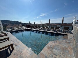 A rooftop pool with clear water, surrounded by lounge chairs and umbrellas, overlooking a cityscape.