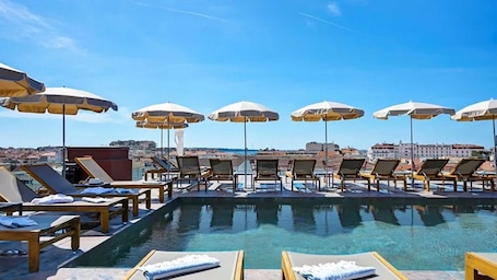 A rooftop pool with sun loungers and umbrellas, overlooking a cityscape.