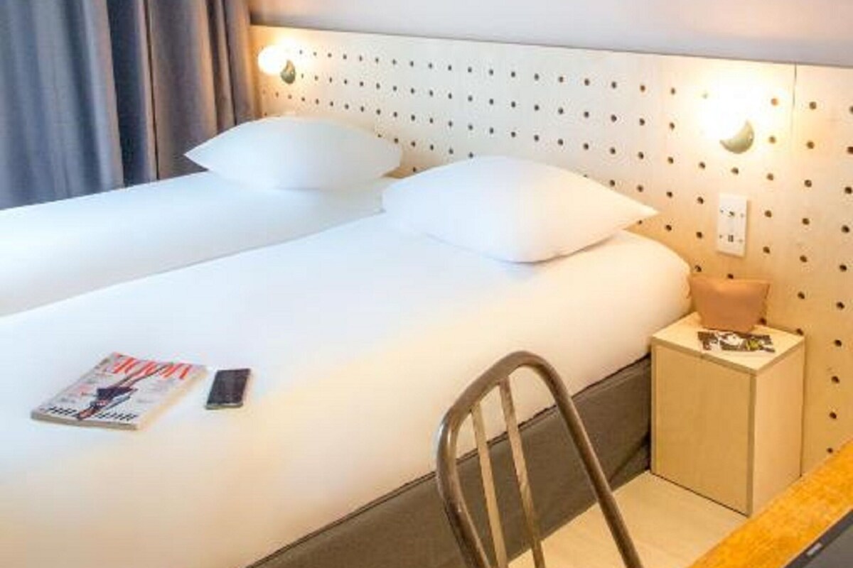 Una habitación de hotel con una cama bien hecha, una mesita de noche con lámpara, una silla y una pequeña mesa con una tableta.