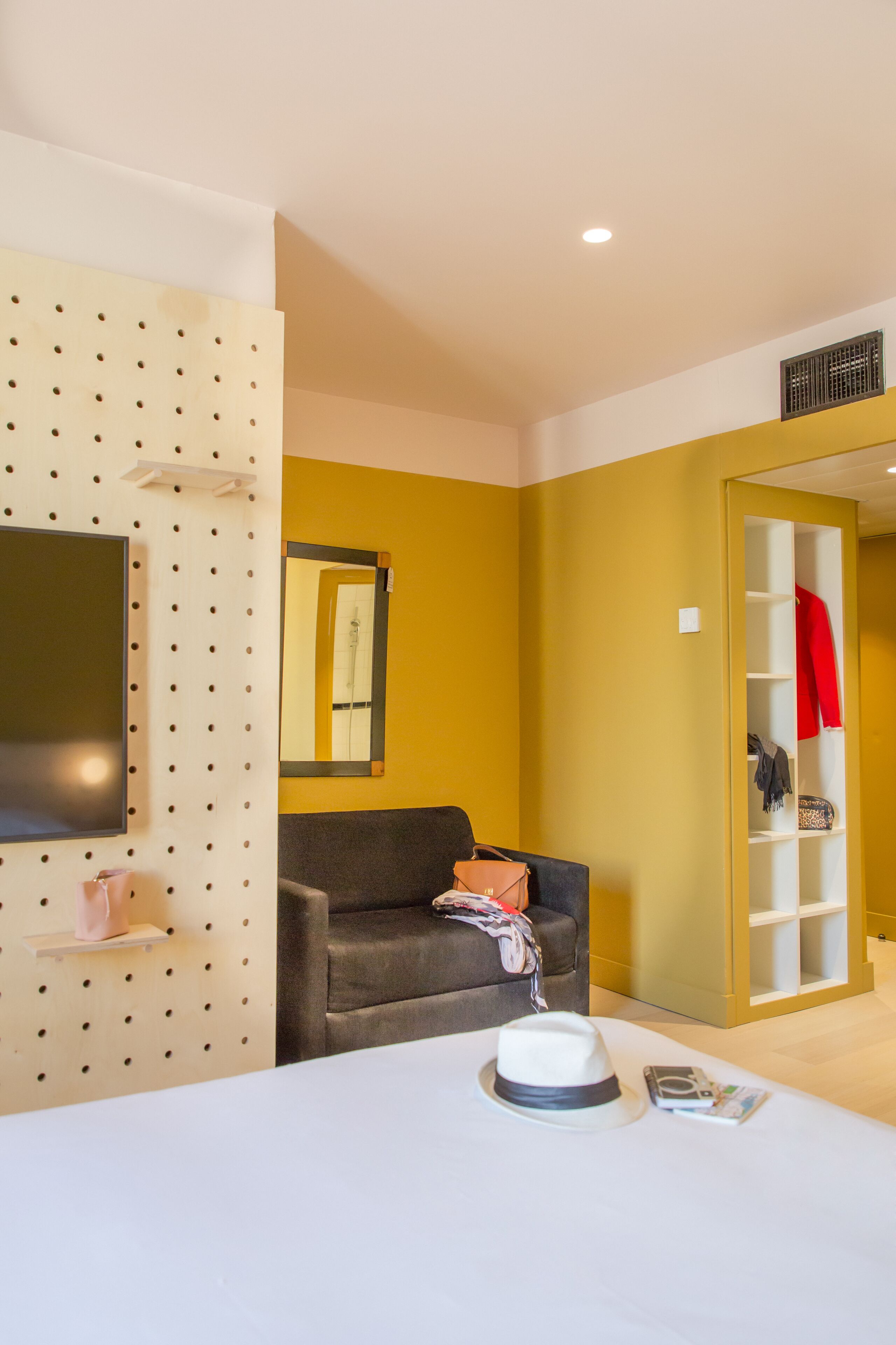 Habitación de hotel con sofá, televisión montada en una pared con un panel perforado, armario con una prenda roja y una mesa con un sombrero y una revista.