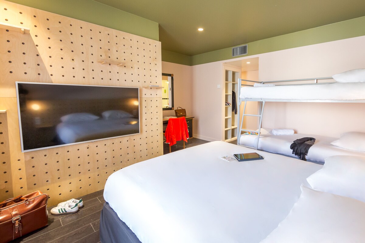 Habitación de hotel con literas, un escritorio con silla, un televisor en la pared y una maleta en el suelo.