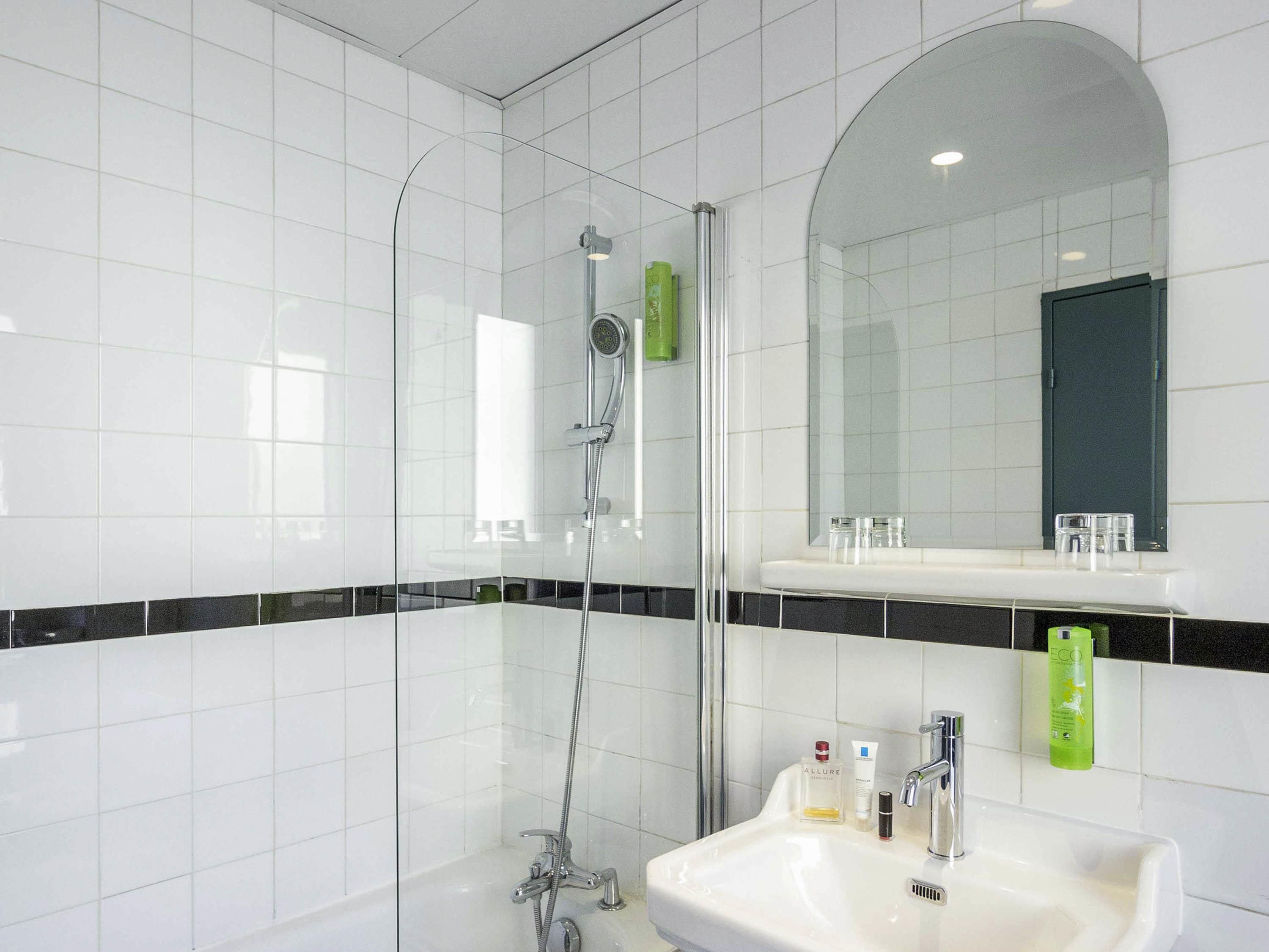 Et moderne badeværelse med hvide fliser, en brusekabine med glasvægge, et vaskebord med spejl og toiletartikler på væggen.