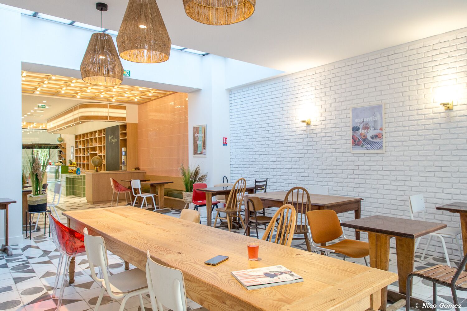 Um café moderno com mesas e cadeiras de madeira, uma parede de tijolos brancos e luminárias de vime suspensas.