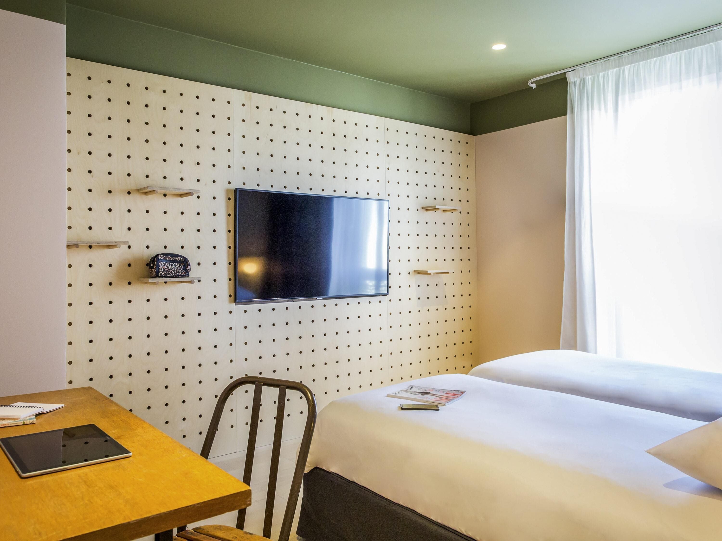 A hotel room with a bed, a desk, a chair, a television mounted on a pegboard wall, and a window with curtains.