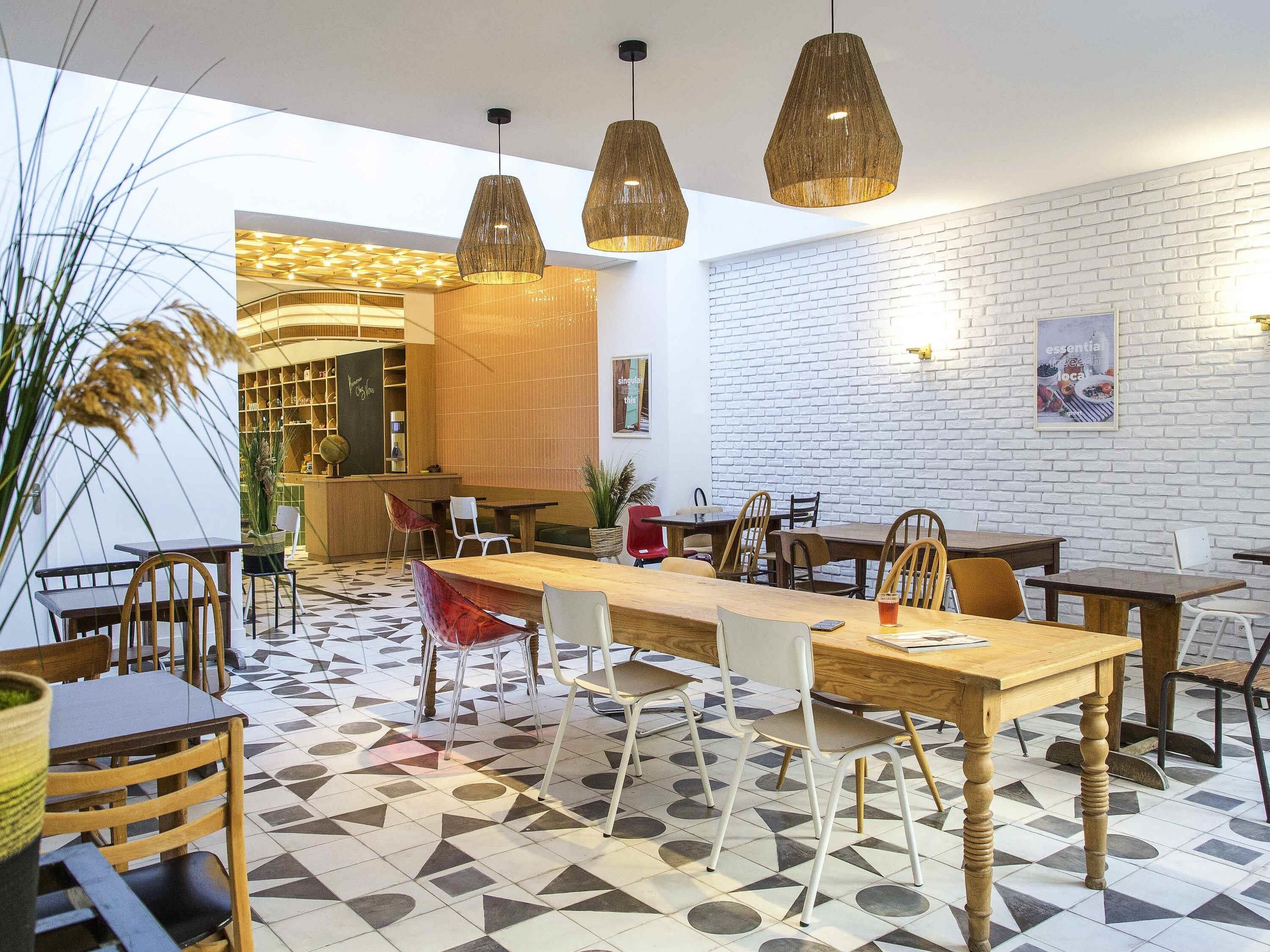 A modern cafe interior with a long wooden table, multiple chairs, hanging pendant lights, and a white brick wall.