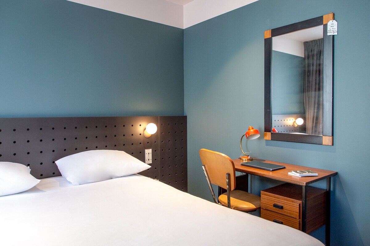 Una habitación de hotel con cama, escritorio, silla, lámpara y espejo.