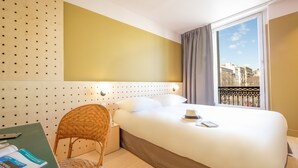 Habitación de hotel con cama, una silla, escritorio con un libro y ventana con vista a edificios.