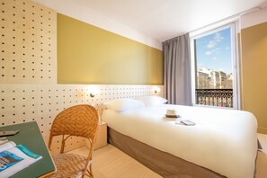 Habitación de hotel con cama, una silla, escritorio con un libro y ventana con vista a edificios.