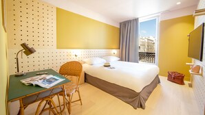Habitación de hotel con cama, escritorio con silla, lámpara y ventana con vistas a edificios.