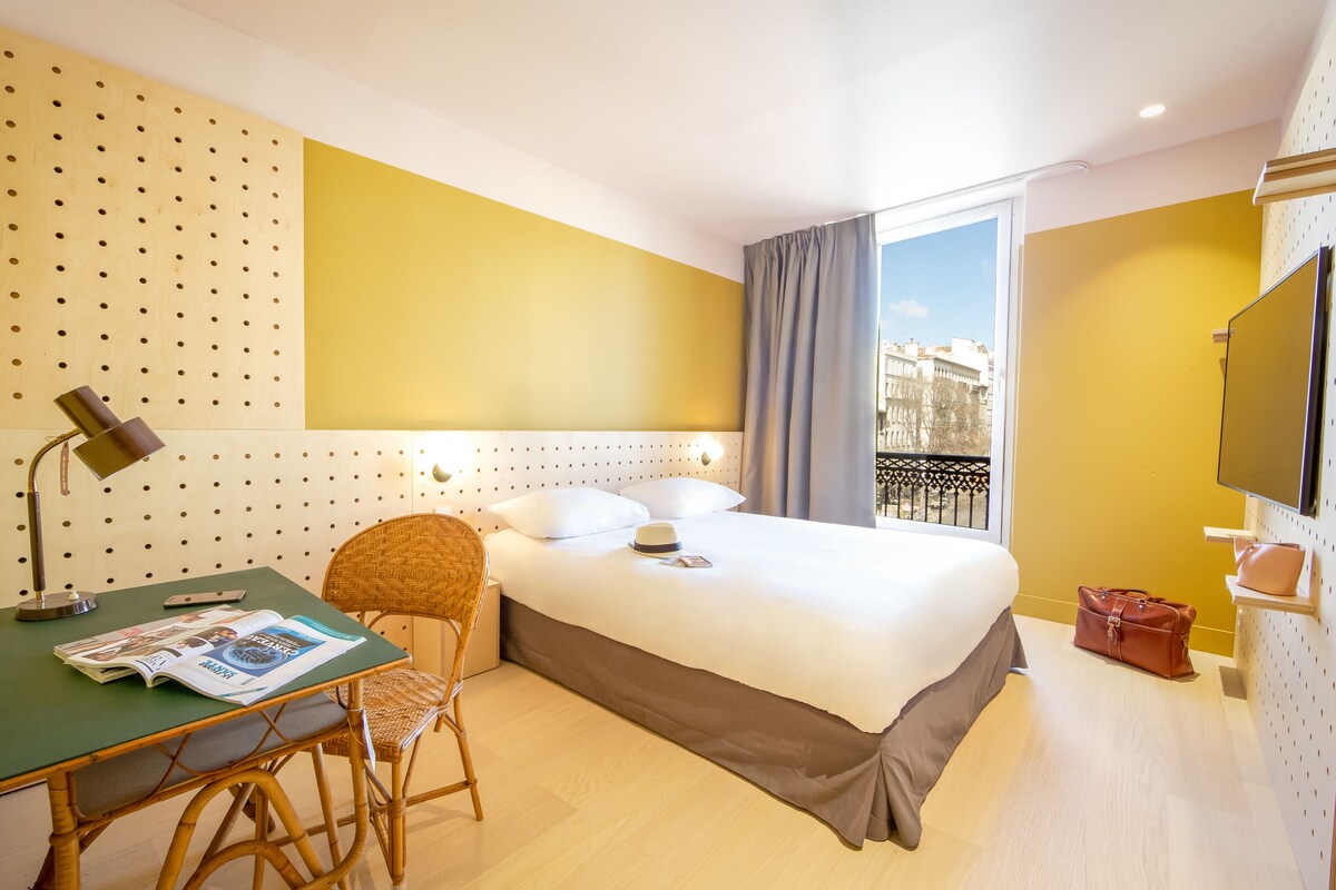Habitación de hotel con cama, escritorio con silla, lámpara y ventana con vistas a edificios.