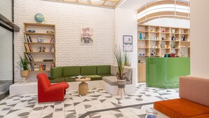 A modern lobby with a green sofa, red armchair, and a glass coffee table. There is a bookshelf with books and decorative items, and a reception desk with a green counter. - Greet Hotel Marseille Centre St Charles (Marseille)