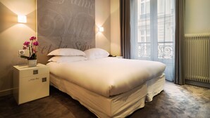 A neatly made bed with white linens, a bedside table with a vase of flowers, and a wall with decorative script. - Hôtel Chambellan Morgane (Paris)
