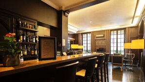 A bar area with a long counter, multiple bar stools, and a shelf with various bottles. There is a potted plant on the counter, and the room has large windows with curtains. - Hôtel Chambellan Morgane (Paris)