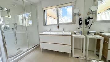Une salle de bain moderne dotée d’une douche à parois de verre, d’un grand meuble-lavabo avec miroir et d’une fenêtre laissant entrer la lumière naturelle.