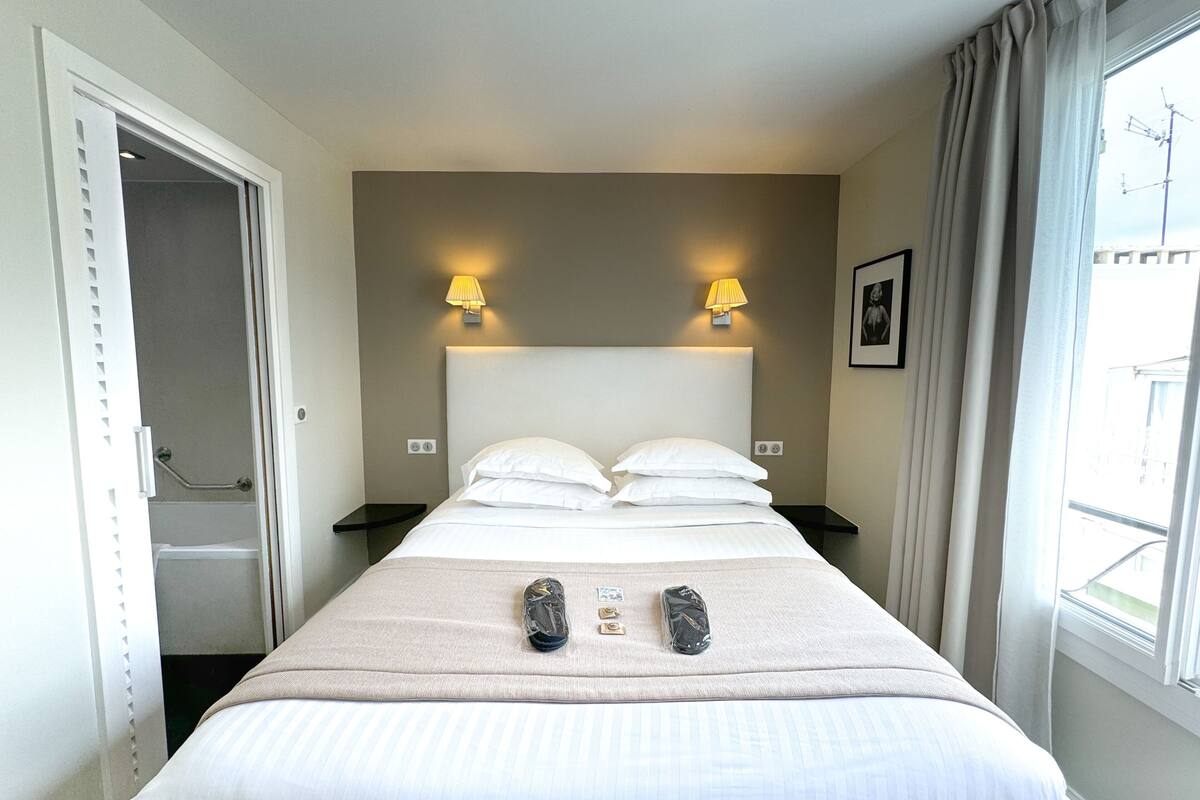A hotel room with a bed, two bedside lamps, a window with curtains, a framed picture on the wall, and a door leading to a bathroom.