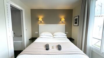 Habitación de hotel con cama, dos lámparas de noche, ventana con cortinas, un cuadro en la pared y puerta que da a un baño.