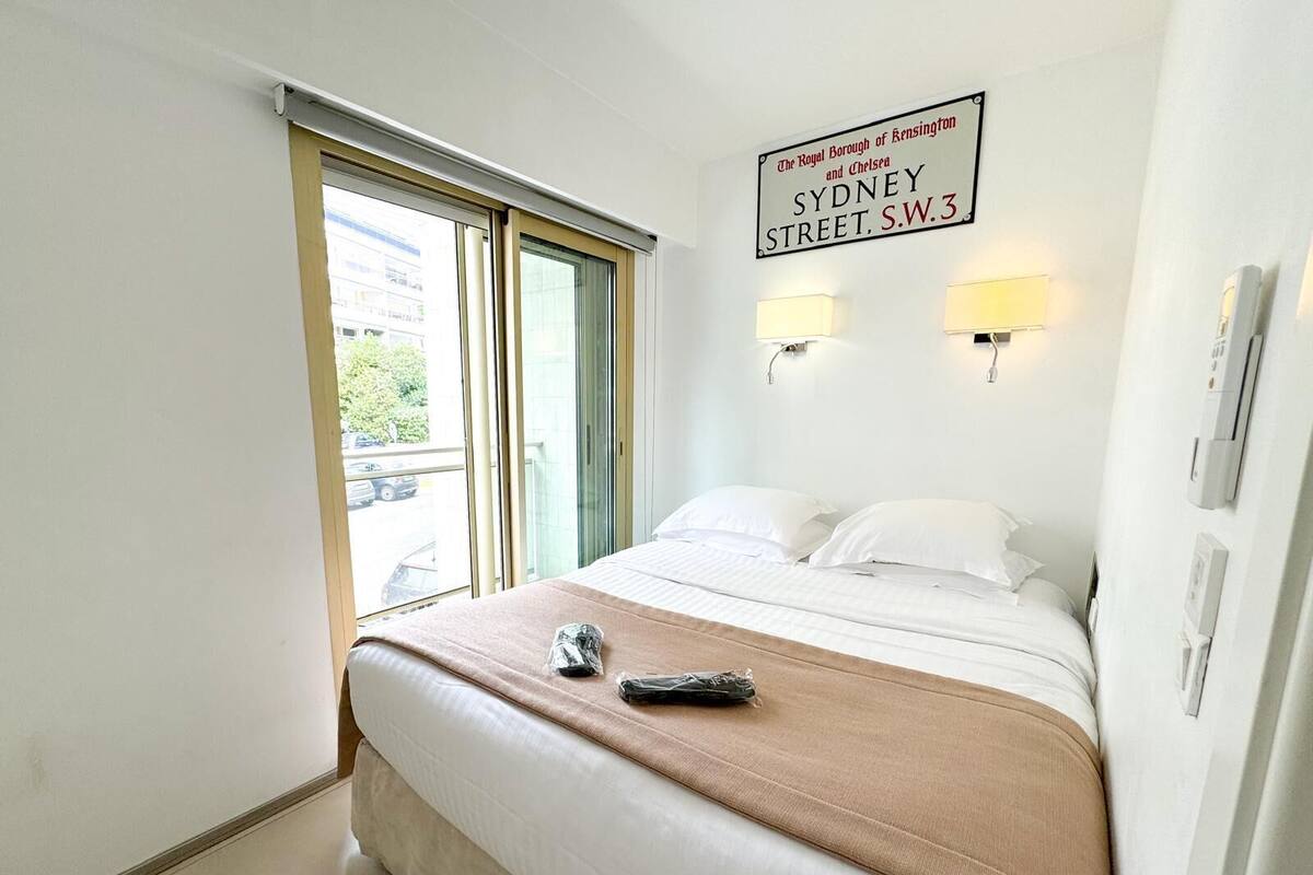 A hotel room with a bed, a window, a sign indicating the location, and a wall-mounted light fixture.