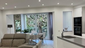 A modern living room with a sofa, dining table, and chairs, a kitchen with a sink and cabinets, and a large window with a view of greenery.