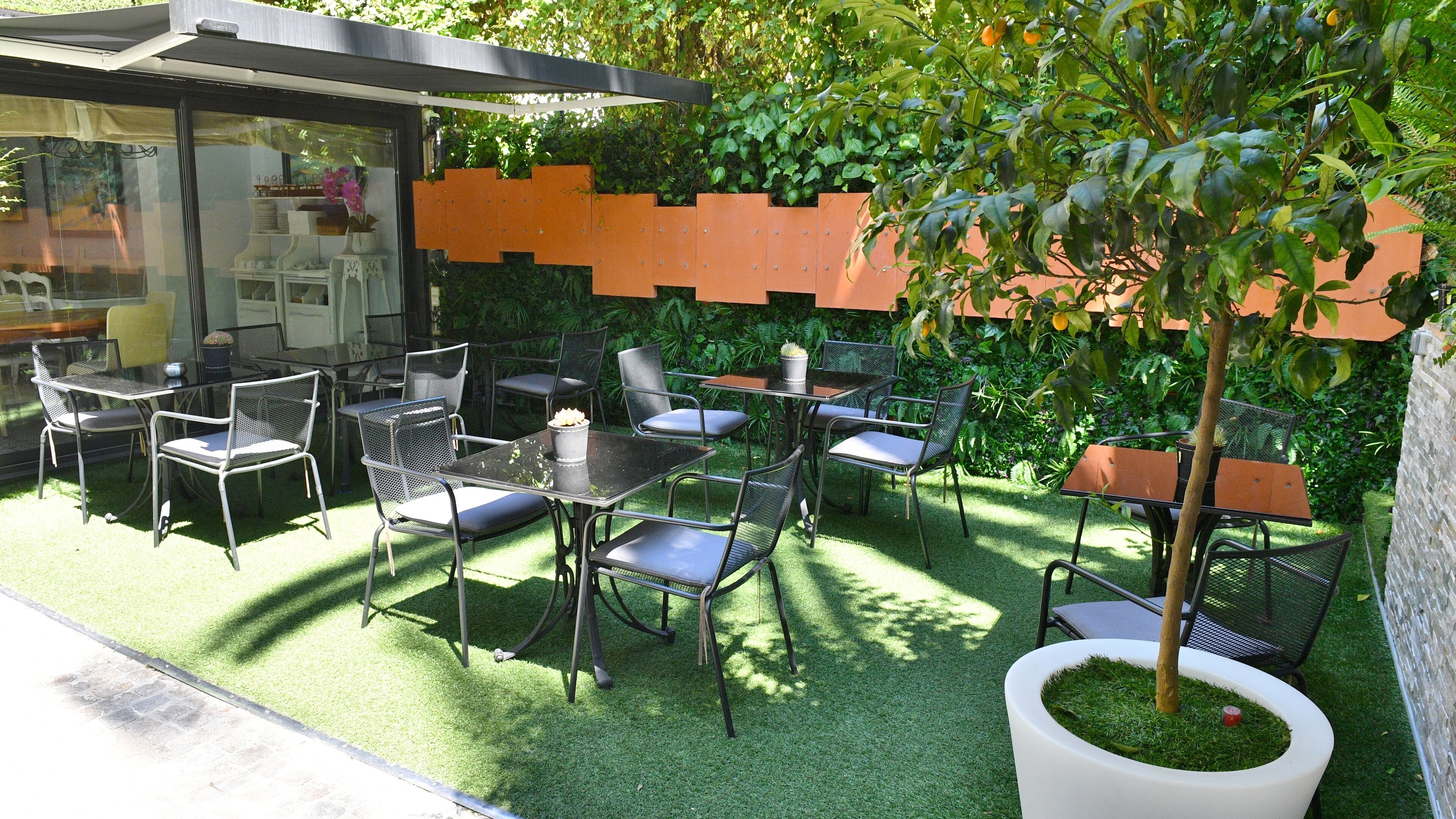 Un patio al aire libre con mesas y sillas, rodeado de follaje y un cerramiento de vidrio.