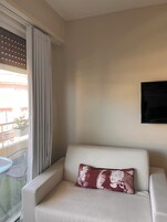 Een witte bank met een rood kussen, een aan de muur bevestigde televisie en een raam met gordijnen.