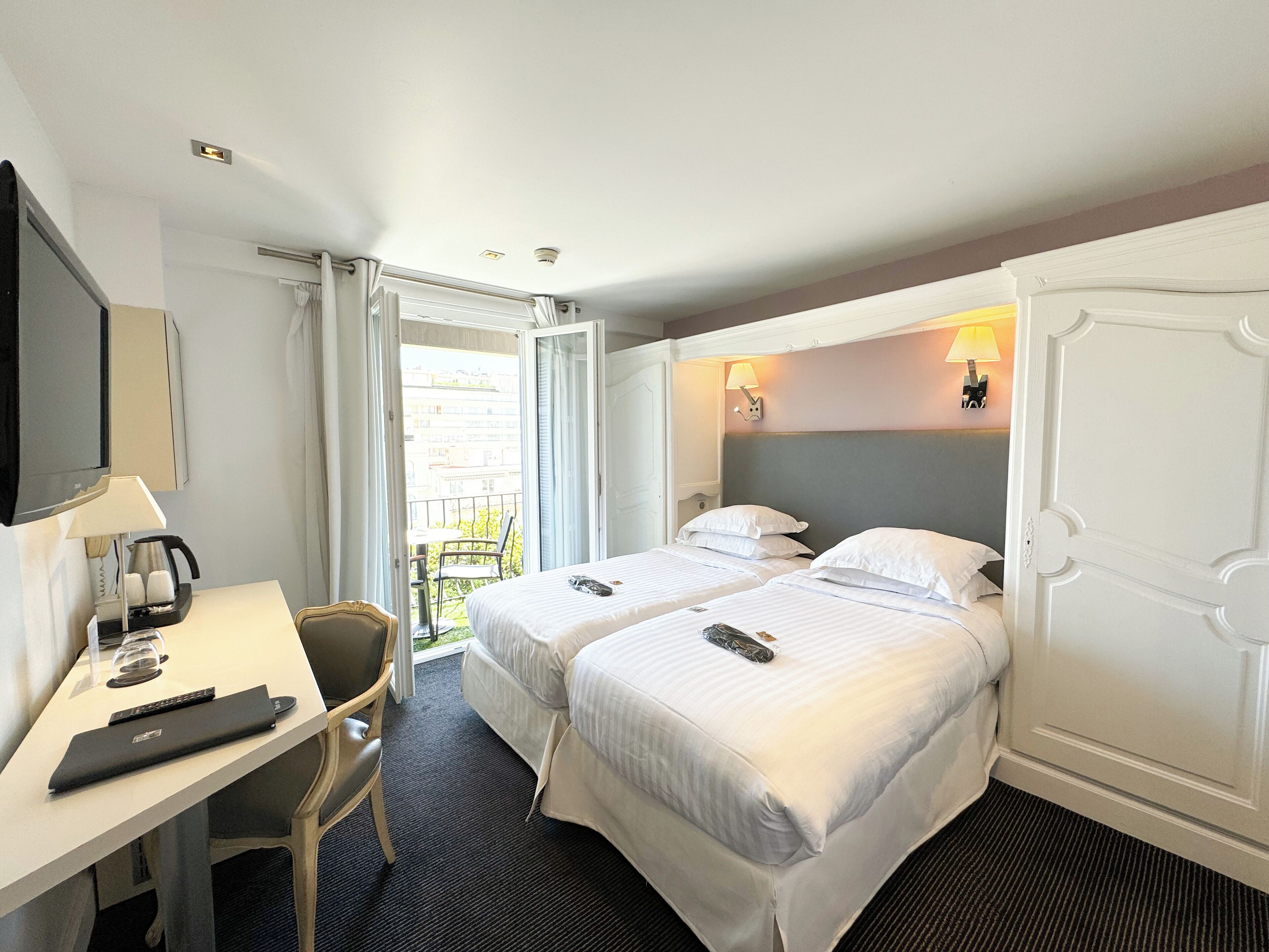 A hotel room with two beds, a desk with a lamp, a chair, and a view of the outside balcony.