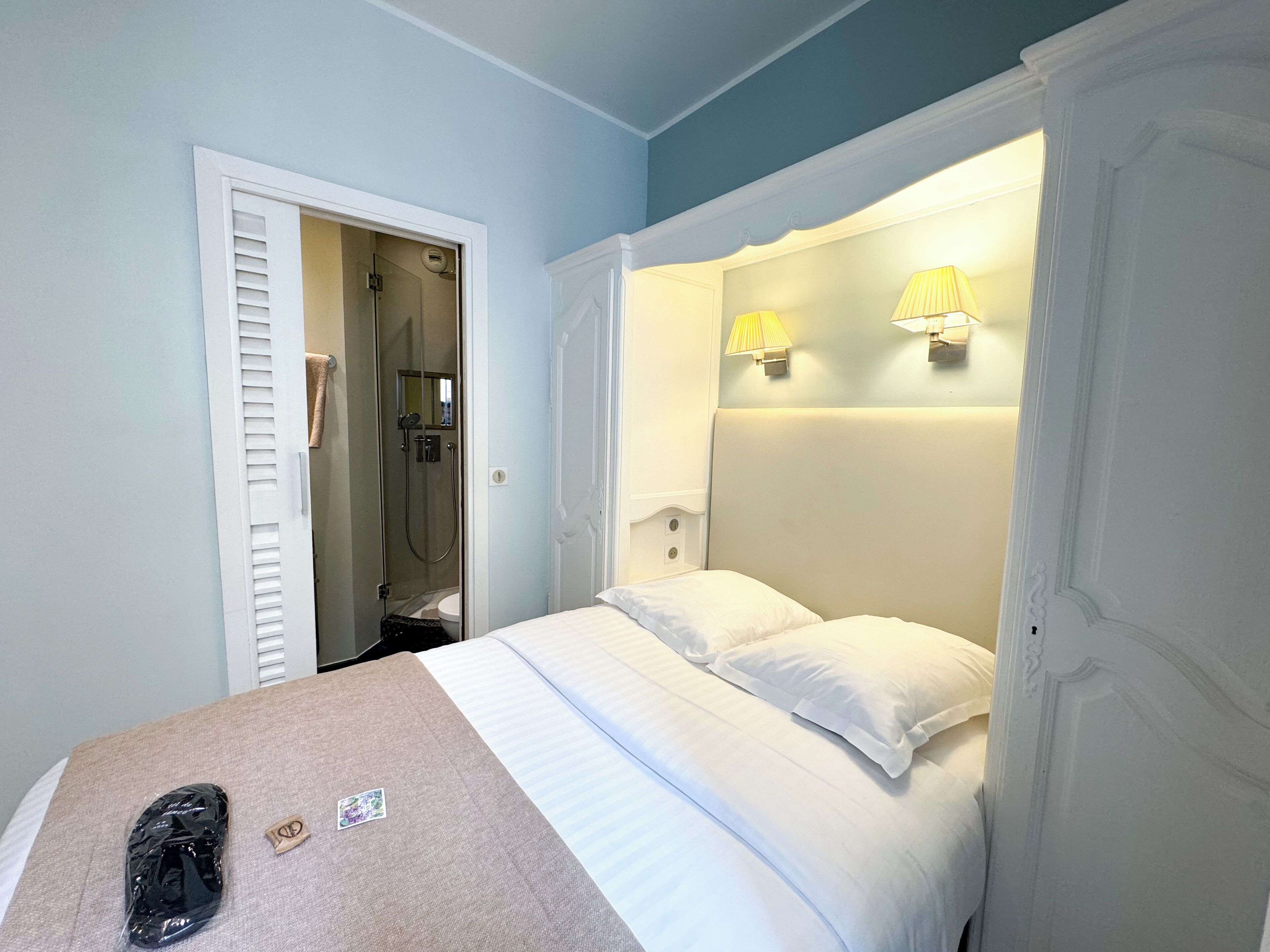 A bedroom with a bed, a bathroom with a shower, and two wall-mounted lamps.