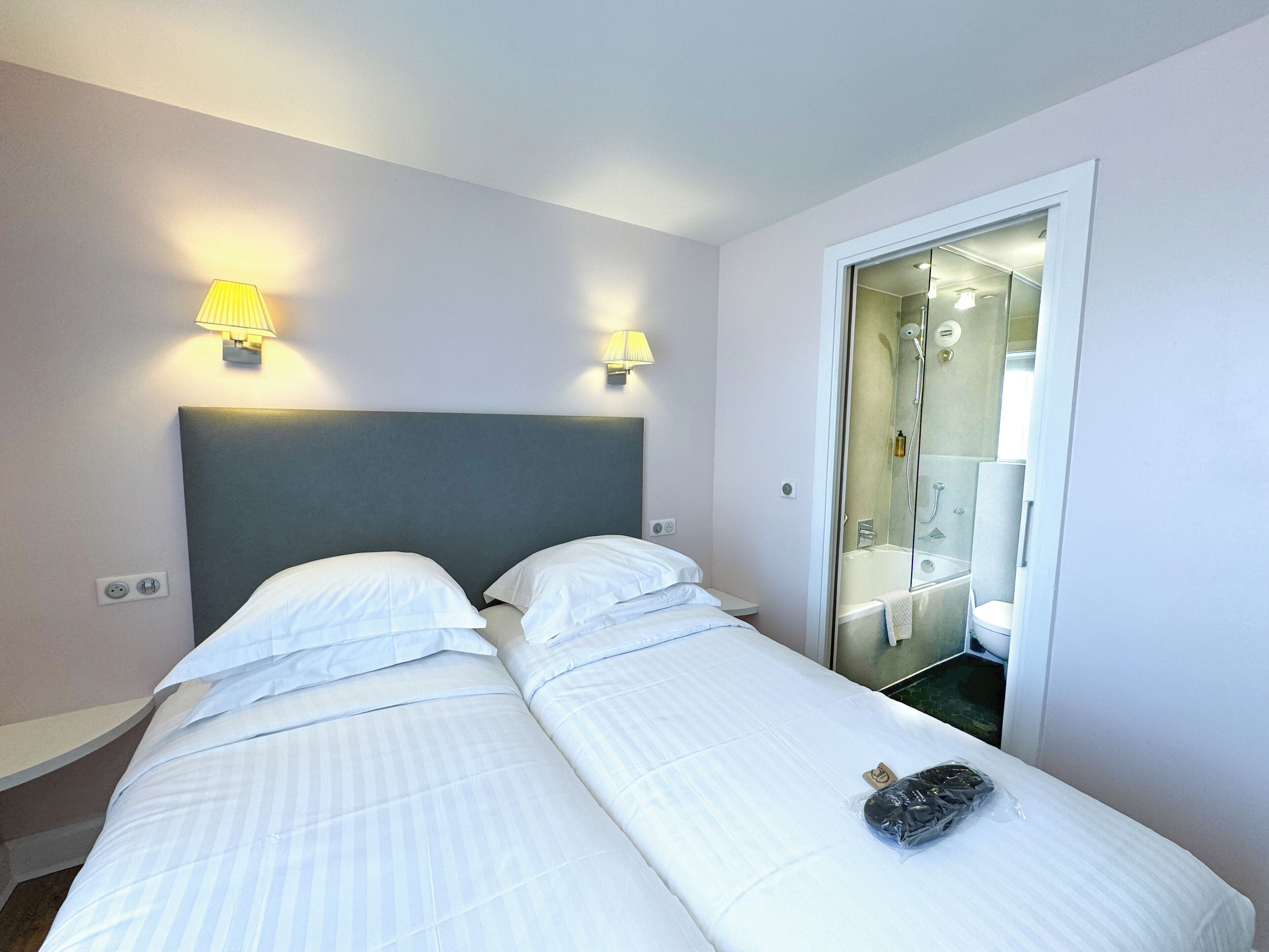 Quarto de hotel com duas camas, cabeceira cinza e duas luminárias fixadas na parede.