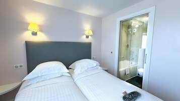 Quarto de hotel com duas camas, cabeceira cinza e duas luminárias fixadas na parede.