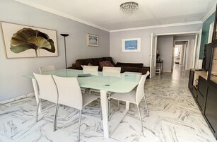 Un comedor con mesa de vidrio y sillas blancas, un sofá oscuro y una araña.