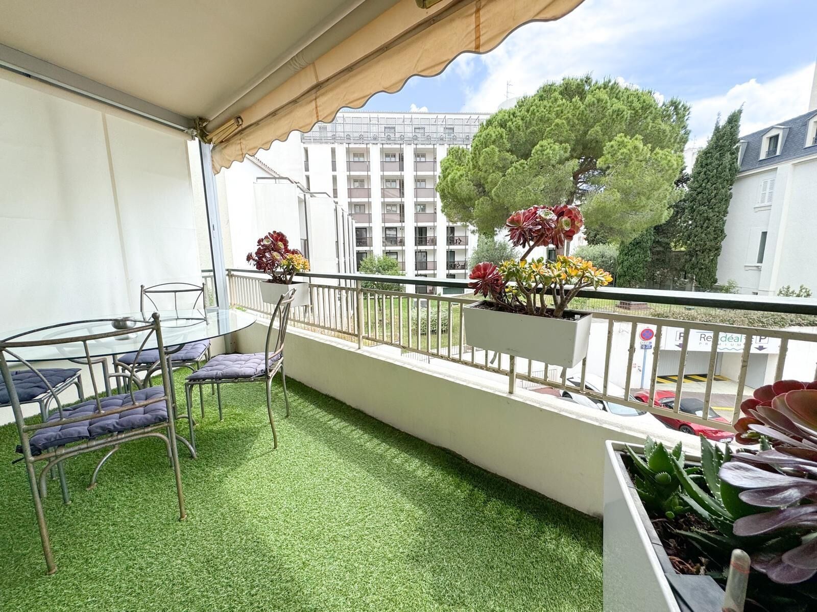 Un balcon avec un revêtement de gazon synthétique, une table en verre, des chaises en métal et des plantes en pot.