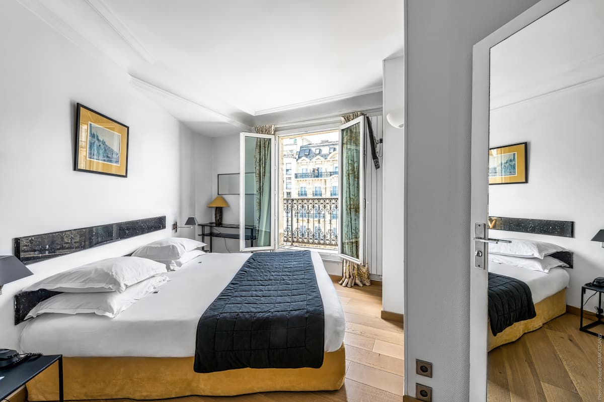 A modern hotel room with two beds, a large window with a view of buildings, and a balcony.