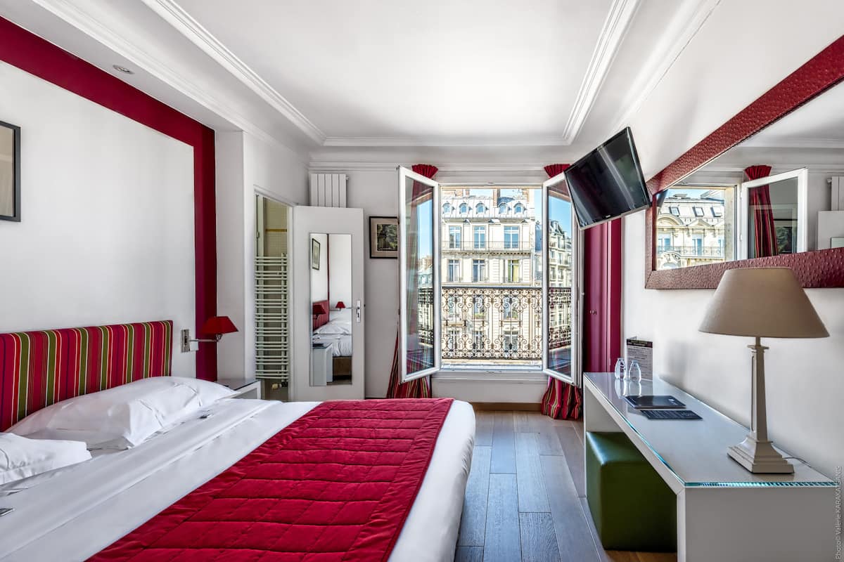 A hotel room with a bed, a desk, a TV, and a window with a view of buildings.