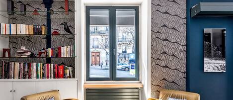 A modern living room with a large bookshelf, two leather armchairs, a wooden coffee table, and a view of the street through a large window.