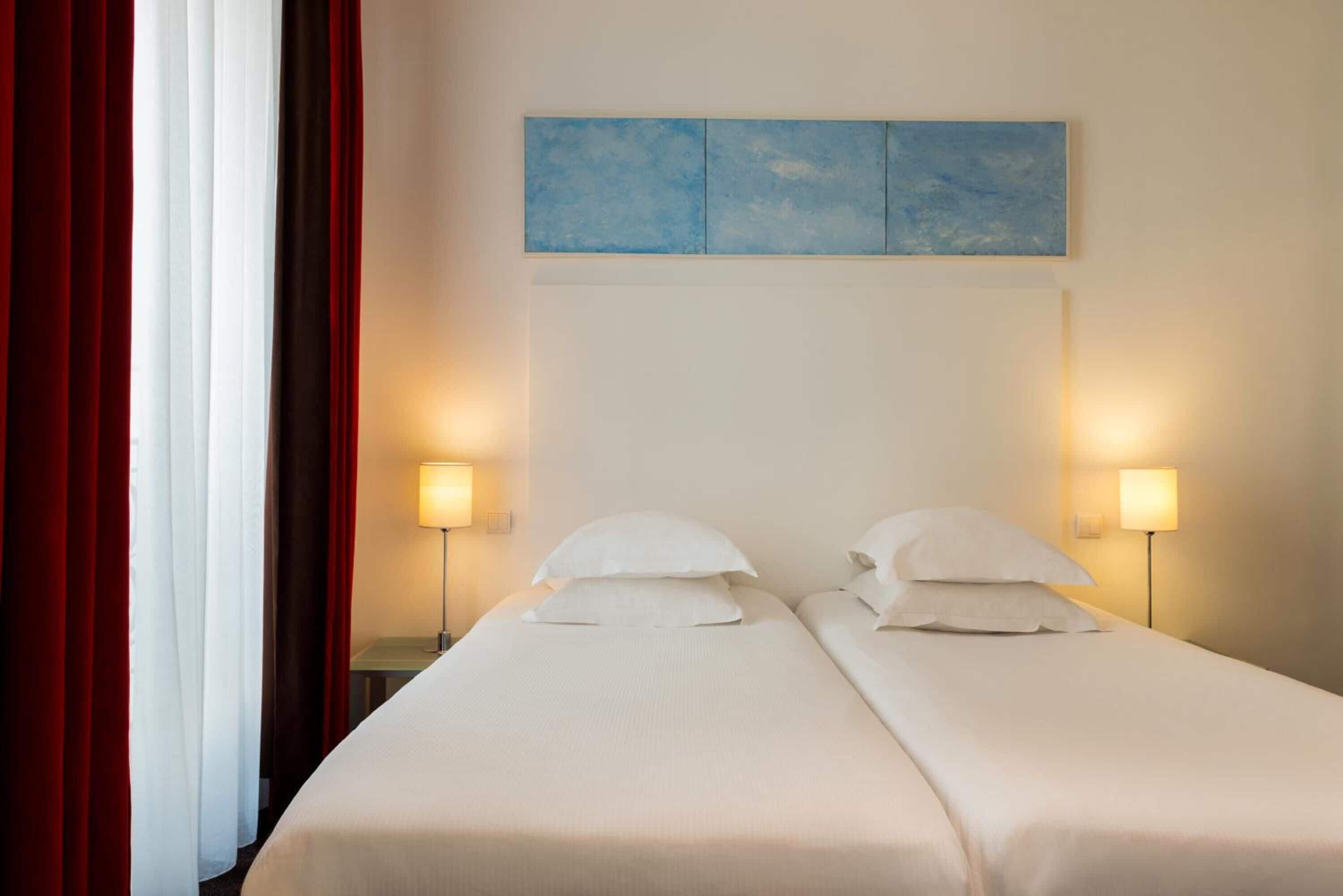 Habitación de hotel con dos camas, cada una con ropa blanca de cama y almohadas, un cuadro abstracto azul sobre las camas y dos lámparas de noche.
