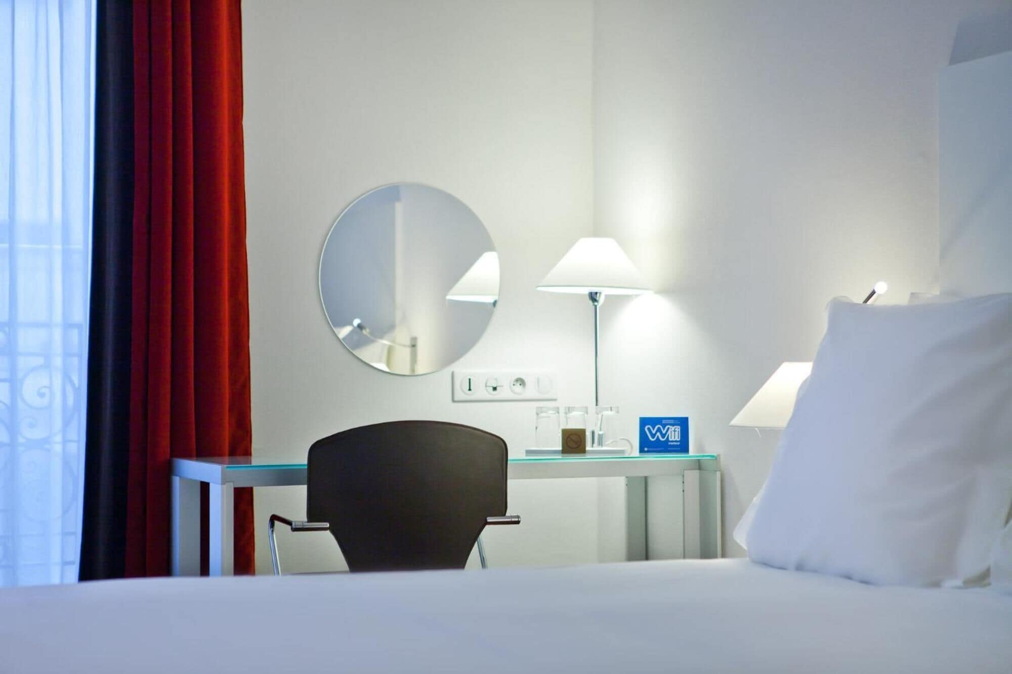 Una habitación de hotel con cama, escritorio, silla, espejo redondo, lámpara y cortina roja.