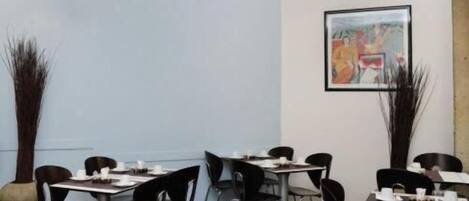 Un comedor con mesas y sillas, un cuadro en la pared y altas plantas decorativas.