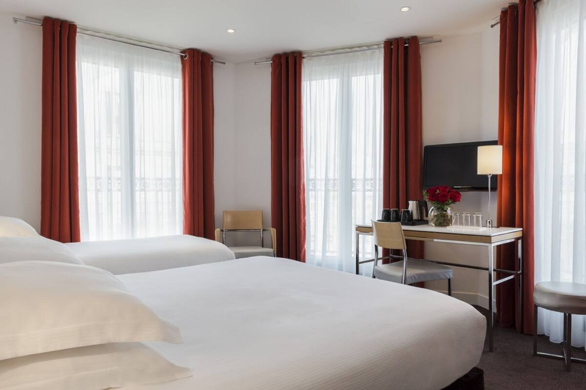 A hotel room with two beds, a desk, a chair, a TV, and a lamp.