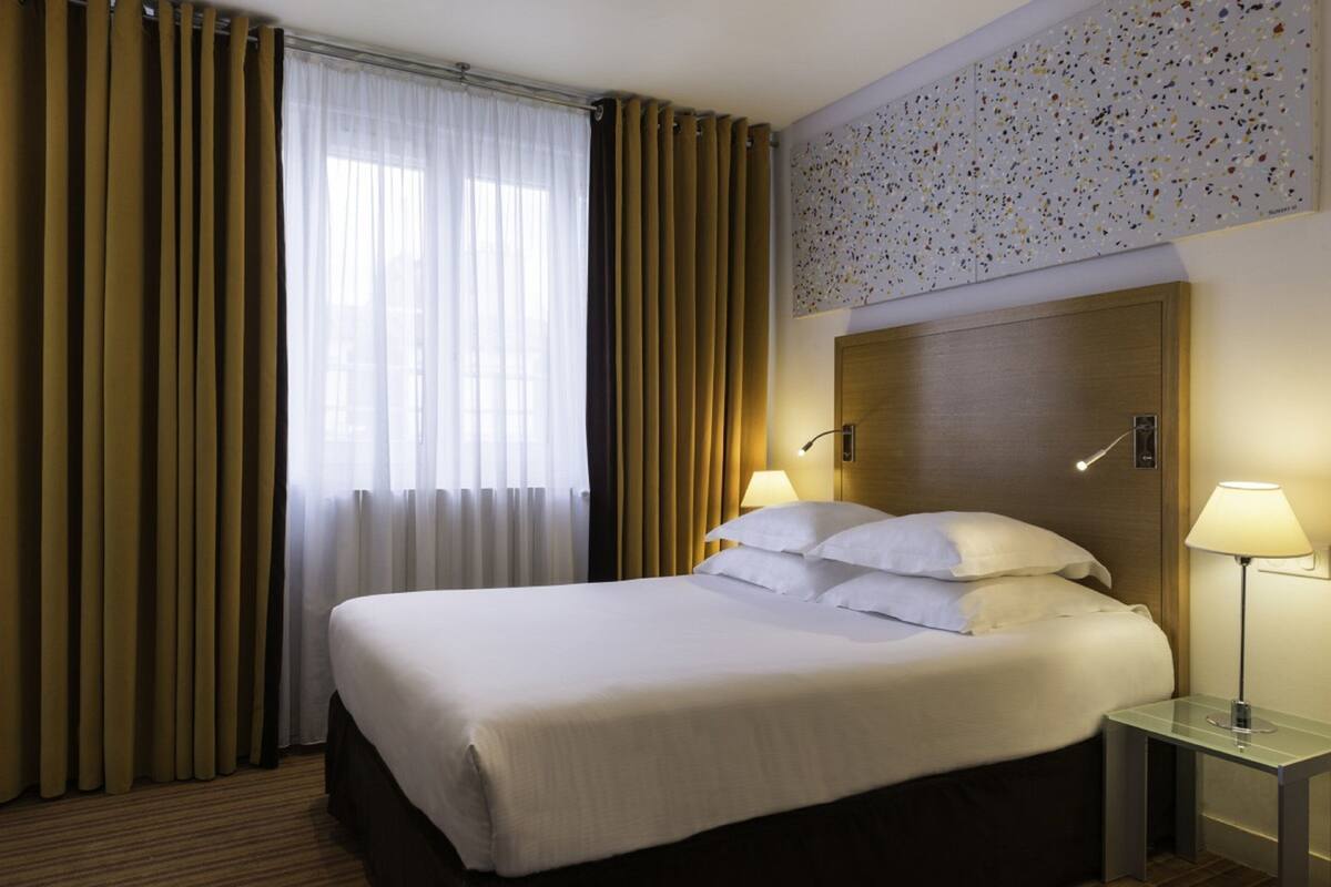 A hotel room with a large bed, two bedside lamps, a headboard, and a window with curtains.