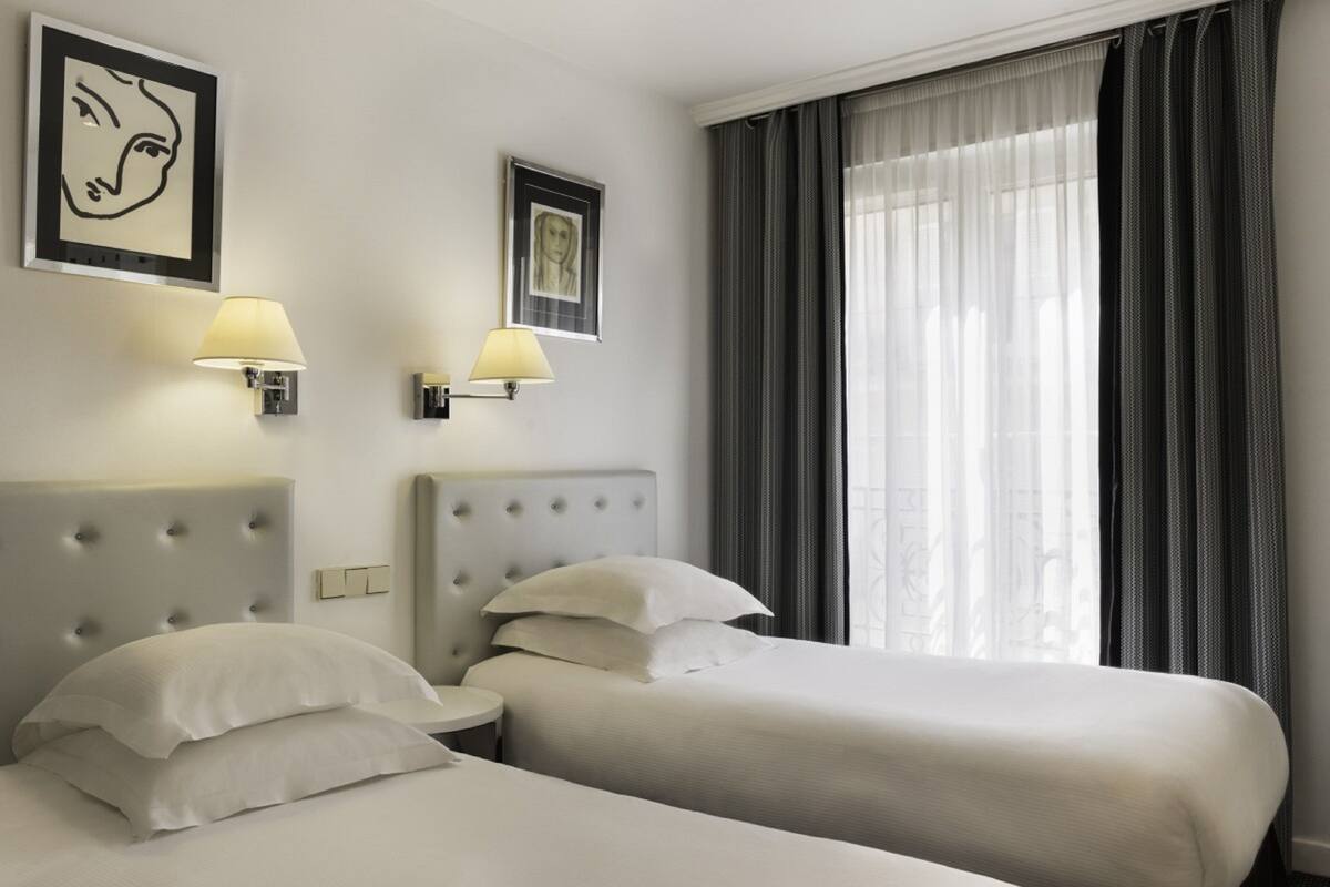 A hotel room with two beds, a large window with curtains, and wall-mounted lamps.