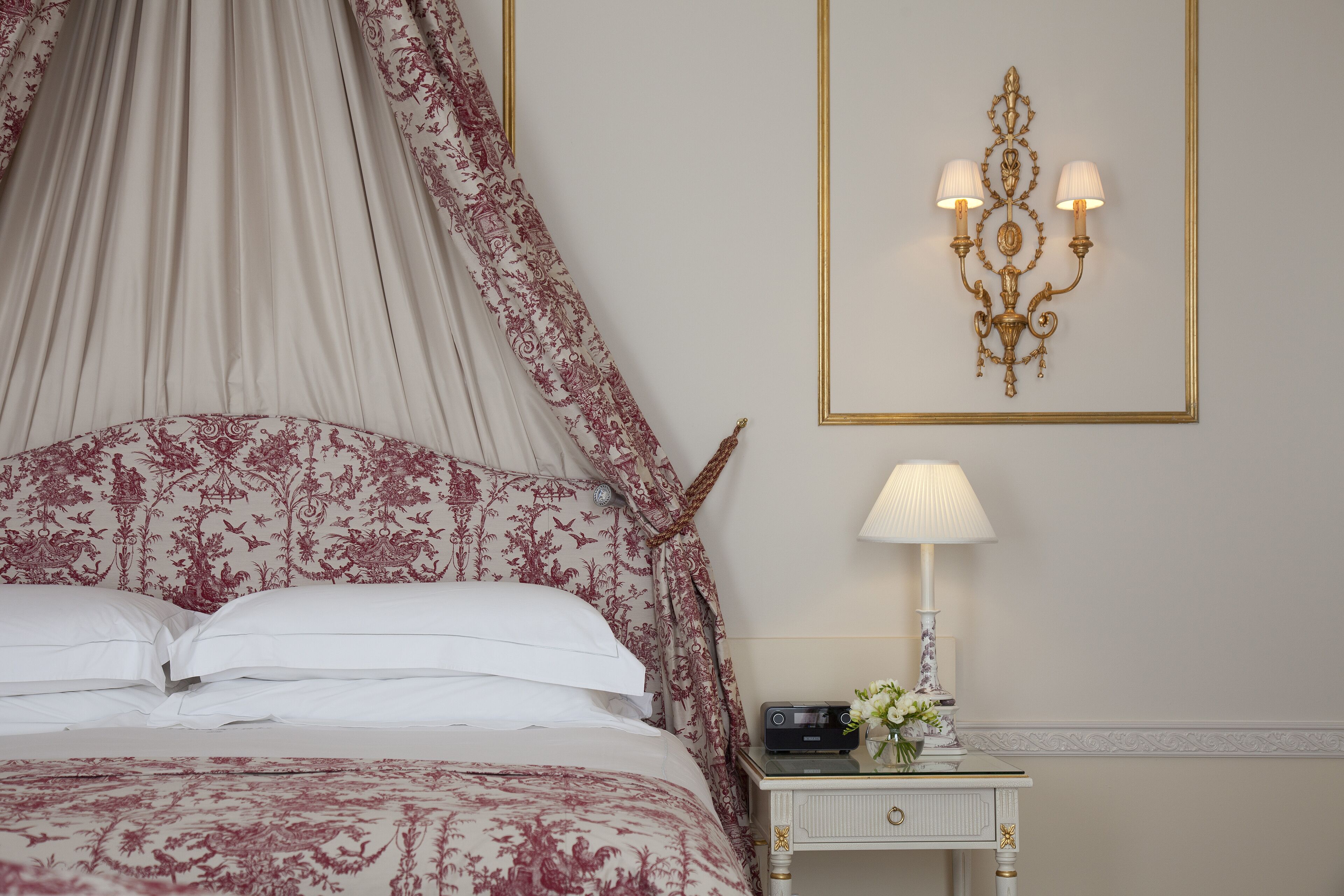 A bedroom with a canopy bed, a bedside table with a lamp, and a wall-mounted light fixture.