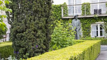 A statue of a man in a suit and hat, a neatly trimmed hedge, and a building with a balcony.