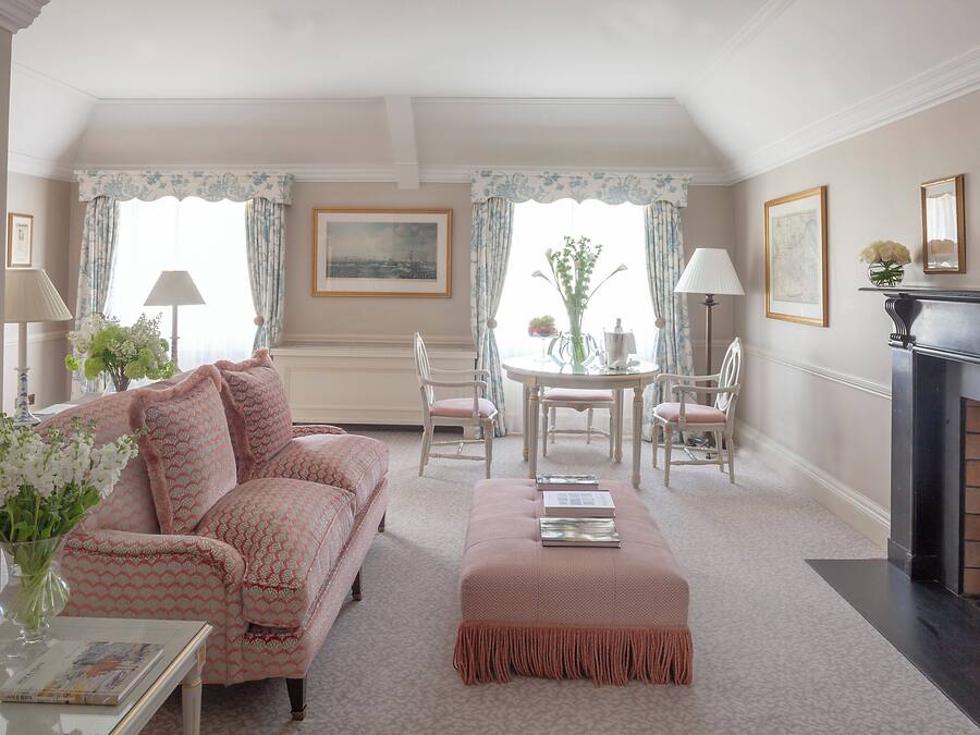 A cozy living room with a sofa, ottoman, and a fireplace. There is a dining table with chairs and a vase of flowers.